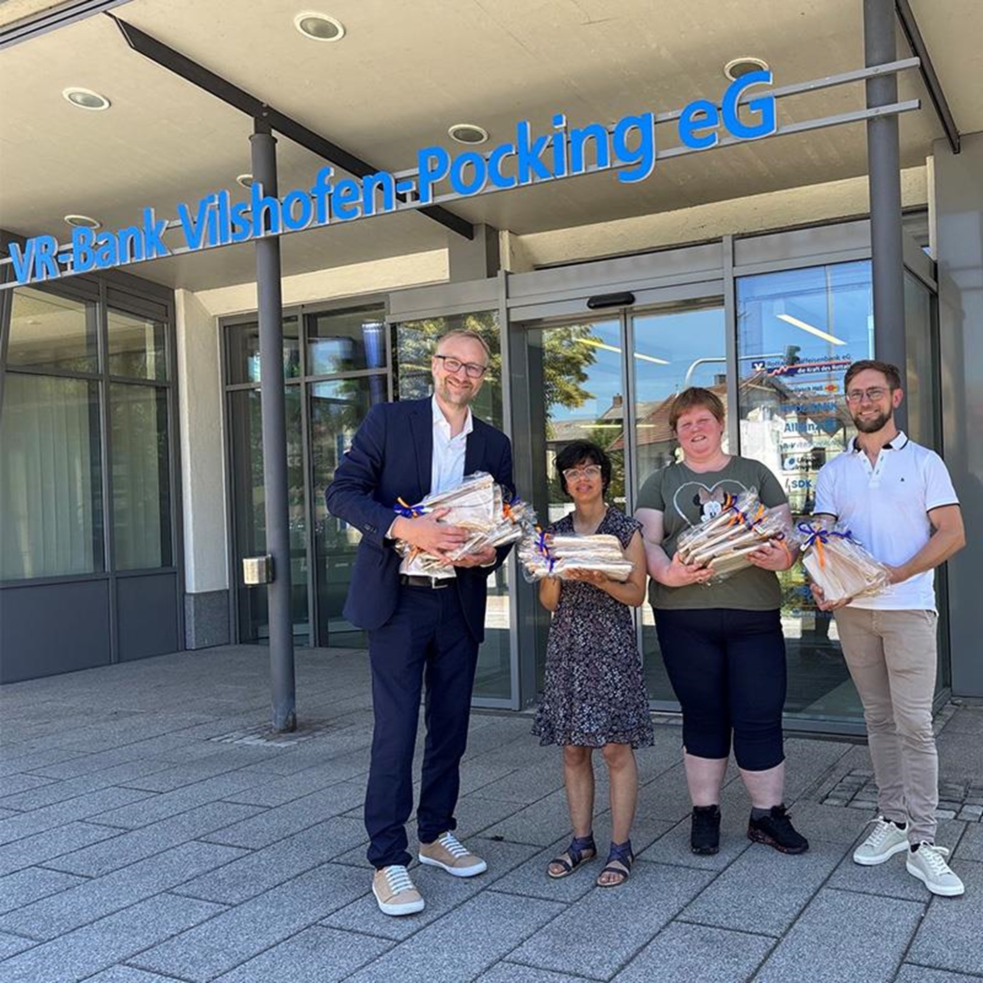 vor einem Gebäude stehen zwei Männer und zwei junge Frauen welche als Geschenk verpackte Holzlöffel in der Hand halten